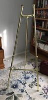 Full view of the gold-colored metal display easel standing on a floral-patterned carpet, showing three-legged structure and top horizontal support.