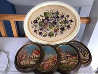 Five framed needlepoint art pieces arranged on a table, one large oval floral design in white frame and four smaller oval countryside scenes in dark frames.