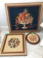 Three needlepoint framed artworks arranged on floor including large fruit still life, medium floral bouquet, and small oval floral piece