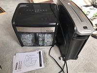 Two paper shredders side by side on carpeted floor, including Staples Mailmate shredder with shredded paper visible inside bin and user manual placed beside it.