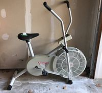 Stationary bike side view showing white frame, fan housing, pedals, seat and elliptical handlebars.