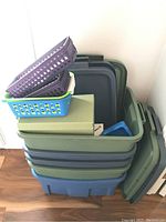 Stacked green and blue plastic storage totes with some lids leaning beside them. Purple and blue ventilated baskets along with a green textured box are placed on top.