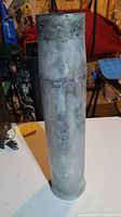Front view of the 105mm tank shell casing standing upright on a table, showing cylindrical shape and metal surface with weathering.