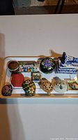 All 11 small decorative boxes arranged on a white tray showing a variety of materials and designs including floral motifs, painted scenes, and a porcelain dog box.