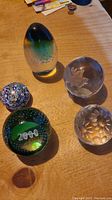 Five glass paperweights displayed on a wooden surface including the Murrina egg-shaped and four round glass paperweights with floral and millefiori designs.