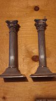 Full view of two metal candlesticks showing the fluted column shafts, trapezoidal bases, and Corinthian capitals.