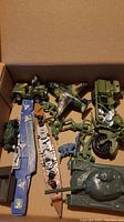 Open box showing green army toy vehicles, plastic toy soldiers, a colorful toy airplane, and two Matchbox ship models.