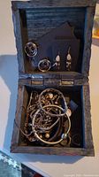 Open wooden carved box showing variety of jewelry inside, including earrings on black card, necklaces and bracelets visible.
