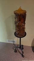 Full view of the tall carved wax candle mounted on black twisted wrought iron style stand with three curled feet.