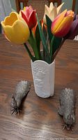 Full lot showing white ceramic vase holding six tulip flowers, two metal peacock figurines on the table around it.