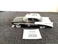 Side profile of black and white collectible highway patrol car model with visible front fender, white roof with red light, and 'Dedicated to Serve' text on rear panel.