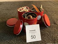 Three red tins containing assorted costume jewelry including faux pearl necklaces, charm bracelets, and colorful bangles, with lids off showing contents.