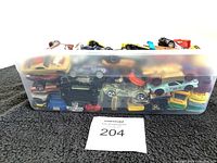 Wide angle shot of plastic bin filled with numerous small toy cars stacked inside, showing different colors and styles visible on top.
