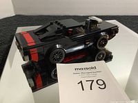 Black Lego muscle car model with red stripe details, angled side view showing rear and roof detail alongside MaxSold card