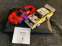Red Lego sports car and white/yellow Audi Team rally car on display board with auction tag visible.