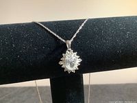 Close-up of silver necklace pendant showing oval central clear gemstone surrounded by small round clear gemstones, necklace draped on black velvet stand.