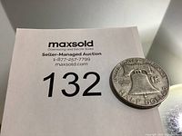 Photo of US half dollar coin displaying the Liberty Bell and engraved text showing denomination and the words 'United States of America'. Coin positioned next to auction lot number paper.