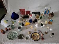Overview of Avon collectible bottles and various souvenir items including mugs, plates, figurines, nesting dolls, and novelty items arranged on a white cloth.