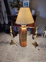 Full view of the copper fire extinguisher lamp with beige lampshade flanked by two matching brass and glass candlestick lamps standing on a tile floor.
