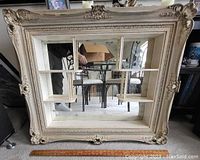 Front view of the large ornate cream-colored framed mirror with three small shelves inside over a split-mirror pane background.