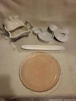 Five serving pieces arranged on a beige surface: covered white porcelain dish on metal stand, two-section white dish with pattern, long white oval tray, and round pink crackle glazed plate.