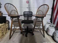 Photo showing two wooden bar stools with spindle backs and one black mesh office chair with caster wheels in a garage setting.