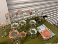 Photo of 10 clear glass lock and seal mason jars of various sizes, 2 lock and seal bottles, and a package of orange rubber seal gaskets on a green cloth surface.