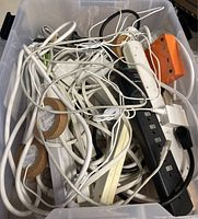 Plastic bin filled with tangled assorted cables and extension cords, mainly white cables with some black and orange items visible.