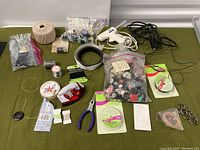 Top-down view of assorted threads, buttons, a white hot glue gun, mini iron, jewelry pliers, metal rings, and various crafting strings on a green surface.