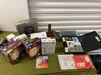 Lot of tools and hardware on green surface including jig saw in box, staple gun with box and staples, packs of LED light bulbs, and furniture anchor kits.