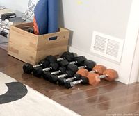 Twelve dumbbells arranged on floor next to wooden box, with black and orange hexagonal heads and chrome handles.