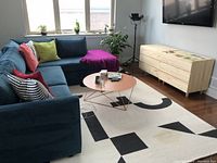 Room view showing the black and white geometric patterned area rug beneath a blue sectional couch, round copper-colored coffee table, and a light wooden media console under a mounted TV.