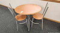 Full view of round wooden bistro table and two matching chairs with silver metal legs and backs.
