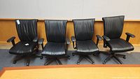 Four black conference chairs aligned in a row showing front and side view, placed on office carpet in front of wood panel wall