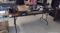 Wide view of the folding table holding various office supplies and organizers. Table is dark brown with metal folding legs. Items include boxes, organizers, and small office supplies.
