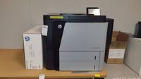 HP LaserJet Enterprise M806 printer on table with toner box at left and cardboard box at right