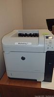 Front angle view showing HP logo and paper output tray with 'Marketing 1' sticker