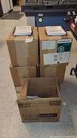 Six sealed cardboard boxes stacked, two spiral-bound notebooks on top of two boxes, each with a white cover and pen attached.