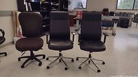 Three wheeled adjustable office chairs arranged side-by-side in room. Two black leather-look executive chairs and one brown fabric chair.