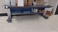 Front view of the long electric lift table showing work surface, shelves underneath with various boxes and materials, and caster wheels.