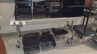 White rectangular office table with metal legs and four caster wheels, surrounded by various black wire organizers on top and underneath.