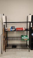 Front view of the 4-tier black wire shelving unit with paper towels on holders and various beverage boxes on shelves.