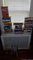 Photo showing multiple stacks of books in varying sizes and conditions arranged on a radiator cover and floor, including both hardcover and softcover editions.