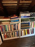 Two white shelving units filled with various hardcover books, mostly historical and educational. Shelving units placed under attic roof rafters.