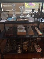 Full view of three-tiered glass candlesticks, metal candlesticks in silver and bronze, boxed candles, and additional candle accessories on black shelving unit.