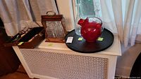 Photo showing two glass decanters in wood carrier, cranberry glass pitcher on wooden lazy Susan, and wood cased travel bar on a white cabinet.