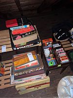 Stack of board games and card decks on wooden slat surface. Includes NFL Strategy, Dominoes, decks of cards, and other boxed games.