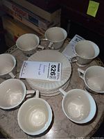 Top view of 8 white porcelain tea cups arranged around 8 matching plates on a tray. Cups have floral design with gold rim.