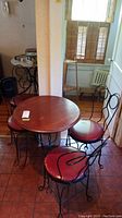Photo showing full round wood topped parlor table and 4 matching metal frame chairs with red upholstered seats in corner next to window