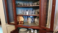 Photo showing three shelves inside a wooden glass door cabinet filled with various glass stemware, pewter platters, copper mugs, and a blue and white ceramic pitcher.